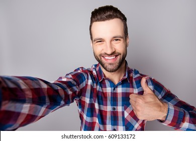 Young handsome excited guy in casual clothing showing thumb up and taking a selfie against gray background.
