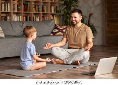 Young Handsome Father In Casual Wear Teaching Little Boy Son How To Meditate While Sitting Together In Lotus Pose On Floor In Living Room At Home, Happy Family Doing Yoga Exercises Online With Laptop