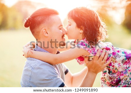 Young handsome father holding, hugging her little cute daughter outdoor in summer park. Fatherhood concept. Dad kissing his pretty cheerful child. Family having fun in park. Male parent loves his baby