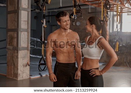 Young handsome fit man and beautiful woman looking to each other and smiling while standing in fitness club