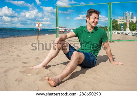 Young handsome fit man wearing green polo and denim shorts enjoys sitting on a sand at the beach on sunny day. Concept of leisure, recreation and well being.