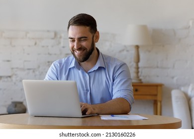 Young handsome freelancer man sit at table using laptop, smiles lead online correspondence to client remotely, share messages enjoy comfort workflow at homeoffice. Modern tech, internet, tech concept