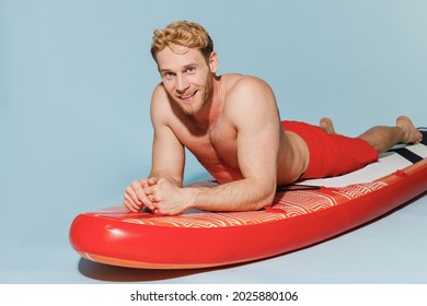 Young handsome fun sexy strong sporty blond man 20s wearing red shorts swimsuit lying on sup board paddle looking camera isolated on pastel blue background. Summer vacation sea rest sun tan concept.