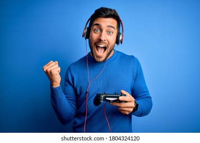 Young handsome gamer man with beard playing video game using joystick and headphones screaming proud and celebrating victory and success very excited, cheering emotion - Powered by Shutterstock