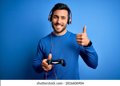 Young handsome gamer man with beard playing video game using joystick and headphones happy with big smile doing ok sign, thumb up with fingers, excellent sign