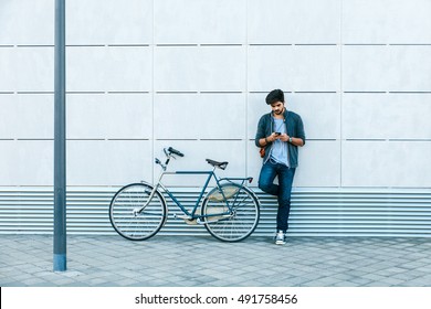 Young handsome guy with a bicycle leaning against the modern wall in the street. He is holding a mobile phone in his hand and listening music.