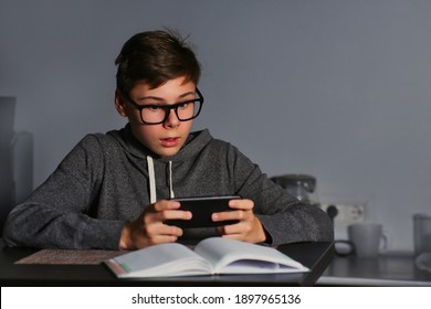 Young Handsome Guy Man In Glasses Looks At The Phone In Surprise, Excitingly Playing Games Online. Communication With Friends On The Internet, A Virtual Date, Relationships. Dark Background, Night.