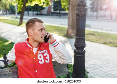 Young handsome guy in a red jacket talking on the phone on a bench in the park