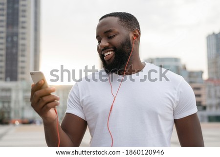 Young handsome guy using mobile phone, communication, looking at digital screen, walking on the street. African American man holding smartphone, listening to music, smiling outdoors