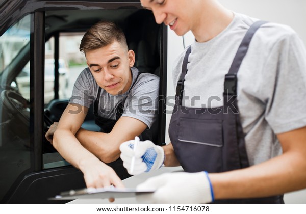 A young\
handsome guy wearing uniform is pointing at the clipboard while\
sitting in the car. Another worker wearing uniform is looking at\
the clipboard. House move, mover\
service.