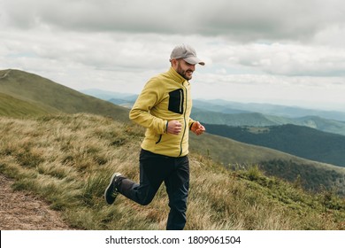 Young handsome happy bearded man in a gray cap, sportswear, yellow jacket and sneakers running in the mountains. Active rest on weekends. Traveling in nature.