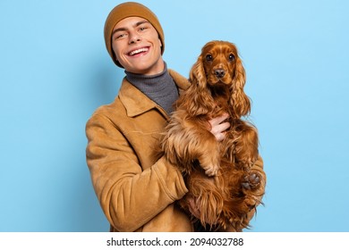 Young Handsome Happy Man, Student Wearing Warm Winter Clothes And Hat Standing With Cute Dog On Blue Studio Background. Concept Of Human Emotions, Facial Expression, Study, Ad, Fashion, Beauty.