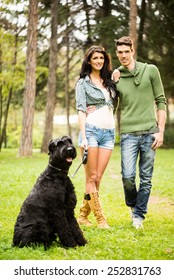 Young Handsome Heterosexual Couple With A Dog, A Black Giant Schnauzer,  Walking Through The Park.