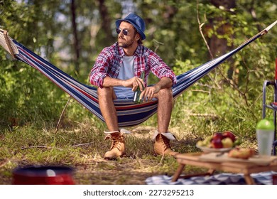 Young handsome hipster male sitting in hammock holding beer. Picnic in forest. Holiday, leisure, lifestyle concept.
