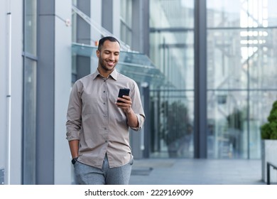 A young handsome Hispanic, African-American man walks down the street near an office center. He is holding a phone in his left hand, his right hand is in his pocket. Uses, reads the news, smiles. - Powered by Shutterstock