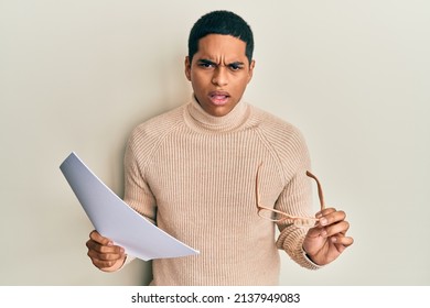 Young Handsome Hispanic Man Holding Blank Paper And Glasses In Shock Face, Looking Skeptical And Sarcastic, Surprised With Open Mouth 