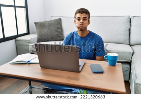 Young handsome hispanic man using laptop sitting on the floor puffing cheeks with funny face. mouth inflated with air, crazy expression. 