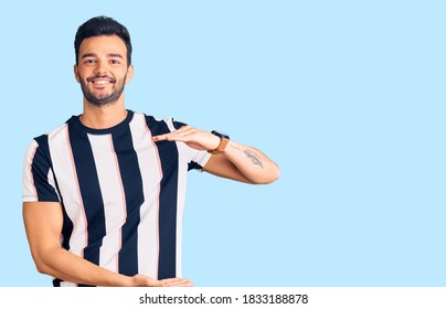 Young Handsome Hispanic Man Wearing Striped Tshirt Gesturing With Hands Showing Big And Large Size Sign, Measure Symbol. Smiling Looking At The Camera. Measuring Concept. 