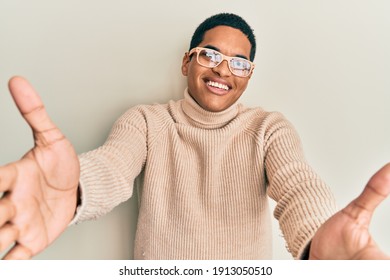 Young Handsome Hispanic Man Wearing Turtleneck Neck Sweater And Glasses Looking At The Camera Smiling With Open Arms For Hug. Cheerful Expression Embracing Happiness. 