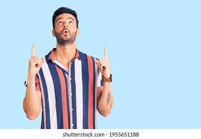Young handsome hispanic man wearing casual  summer shirt amazed and surprised looking up and pointing with fingers and raised arms. 
