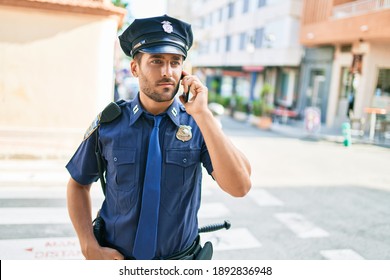 Young Handsome Hispanic Policeman Wearing Police Uniform Smiling Happy. Standing With Smile On Face Having Conversation Talking On The Smartphone At Town Street.