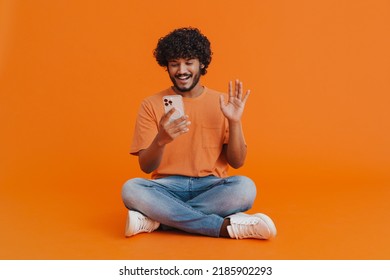 Young Handsome Indian Smiling Man Sitting With Phone In Lotus Pose Holding Video Conference And Waving Over Isolated Orange Background