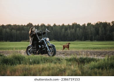 Young handsome long-haired man riding on motorbike at countryside road with his dog runs nearby
