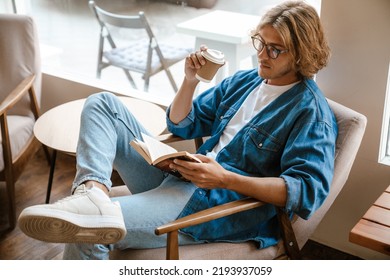 Young handsome long-haired stylish man in glasses reading book while sitting with coffee in armchair in cafe - Powered by Shutterstock