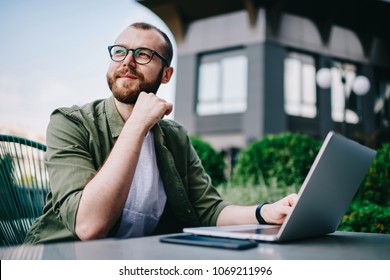 Young handsome male 20 years old in eyeglasses sitting at street cafe during free time with new modern laptop, bearded man freelancer  thoughtfully looking aside and thinking about distance work