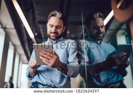Young handsome male with beard in classic formal wear focused on devise standing near glass wall with reflection and looking away