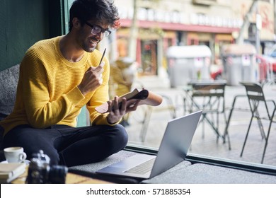 Young Handsome Male Blogger Watching Web Workshop Noting Ideas Into Personal Organizer Starting Working Day With Cup Of Coffee Sitting With Laptop In Cafe Using Free Wireless Connection To Network