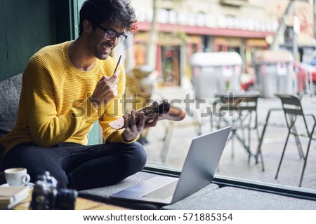 Young handsome male blogger watching web workshop noting ideas into personal organizer starting working day with cup of coffee sitting with laptop in cafe using free wireless connection to network