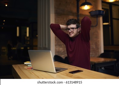 Young handsome male football fan in eyewear and casual wear watching streaming video of match cheering for team feeling disappointed of bad game of players sitting in coworking space with wifi zone