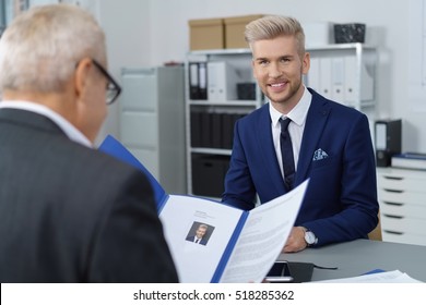 Young Handsome Male Job Seeker In Blue Jacket And Dark Necktie Having An Interview As Boss Looks At His Resume