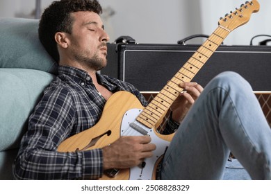 Young handsome male musician playing electric guitar concentrated on the floor of his living room next to the guitar amplifier, playing blues or rock alone - Powered by Shutterstock