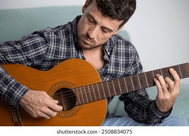 Young handsome male musician playing classical Spanish guitar concentrated on the sofa in the living room of his home, playing blues, flamenco, jazz or rock and roll alone - Powered by Shutterstock