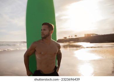 Young handsome male surfer with surfboard posing on sandy beach at sunset, muscular athletic guy standing on seaside and looking aside, waiting for high waves, enjoying surfing and summer vacation - Powered by Shutterstock