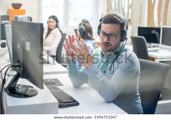 Young handsome male technical support agent trying\
to explain something to a client while using hands-free headset at\
call center