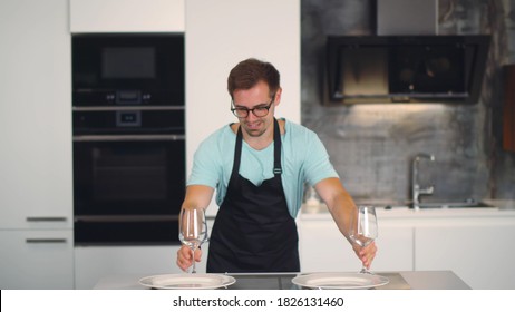 Young Handsome Man In Apron Dancing With Wine Glasses Setting Table For Romantic Dinner In Kitchen. Cheerful Boyfriend Putting Dishware On Table Preparing For Date In Home Kitchen