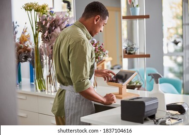 Young Handsome Man In Apron Is Standing At Counter With Tablet And POS Terminal While Selling Bouquets