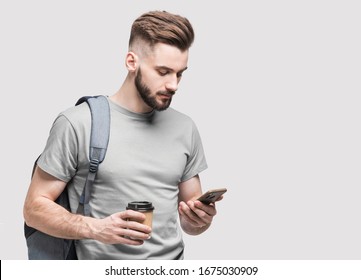 Young Handsome Man With Backpack Using Smart Phone And Holding Coffee Isolated On Gray Background. Serious Student Or Businessman Looking At Mobile Phone, Studio Portrait, Isolated On Gray Background