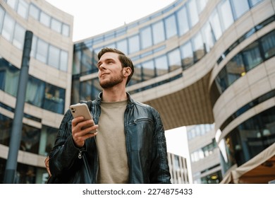 Young handsome man with backpack using mobile phone and looking aside while standing outdoors at city street