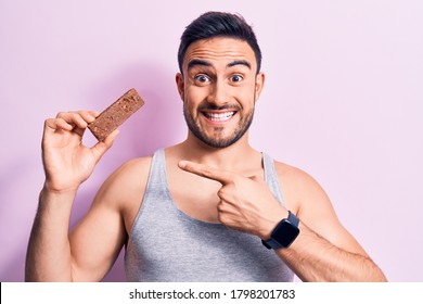 Young Handsome Man With Beard Eating Energy Protein Bar Over Isolated Pink Background Smiling Happy Pointing With Hand And Finger