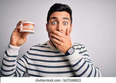 Young Handsome Man With Beard Holding Plastic Denture Teeth Over White Background Shocked Covering Mouth With Hands For Mistake. Secret Concept.