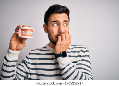 Young handsome man with beard holding plastic denture teeth over white background looking stressed and nervous with hands on mouth biting nails. Anxiety problem.