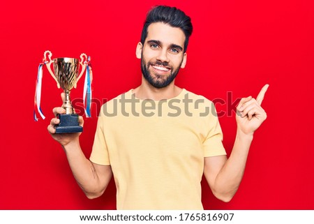Young handsome man with beard holding trophy smiling happy pointing with hand and finger to the side 
