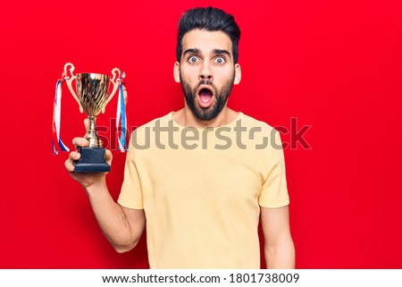 Young handsome man with beard holding trophy scared and amazed with open mouth for surprise, disbelief face 