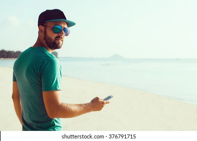 young handsome man with beard and mustache in baseball cap, with a smartphone in hand, hipster style, outdoor portrait, close up,posing on the beach in a t-shirt and denim shorts, the bearded - Powered by Shutterstock