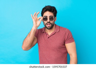 Young handsome man with beard over isolated blue background with glasses and surprised