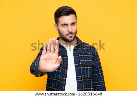 Young handsome man with beard over isolated yellow background making stop gesture